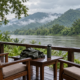 เก้าอี้ไม้ริมแม่น้ำกับภูเขาเขียวในเช้าวันฝนพรำ